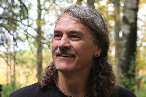 Man with long gray hair, wearing a t-shirt, beaded necklace, and an earring smiles at something to his right.