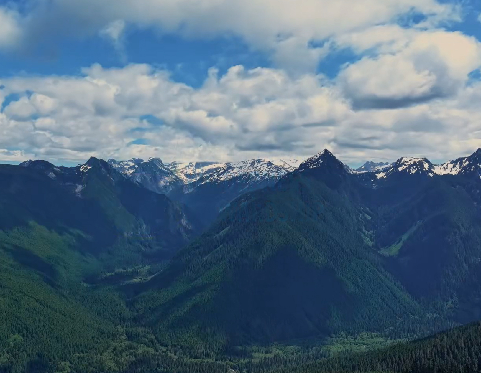 Mountain peaks rise up out of darkly wooded valleys, against a blue sky with scattered clouds.