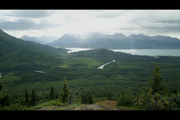 Mountains, green forest, a river and a large body of water, all reflect bright sunlight shining through moody clouds, in what appears to be an Alaskan landscape.