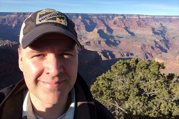 Man wearing ballcap smiles at the camera in front of the Grand Canyon.