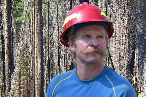 A mustachioed fellow in a red hardhat, holding a clipboard, looks at something slightly overhead, off camera to his left.