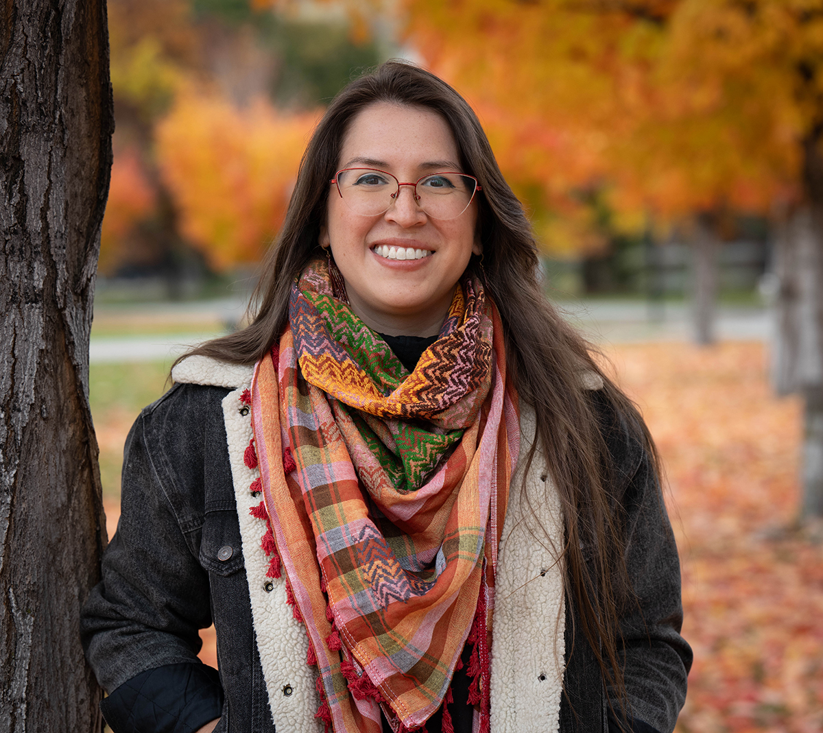 Young woman wearing a jacket, brightly colored scarf, and glasses stands amidst maple trees in full autumn color.  