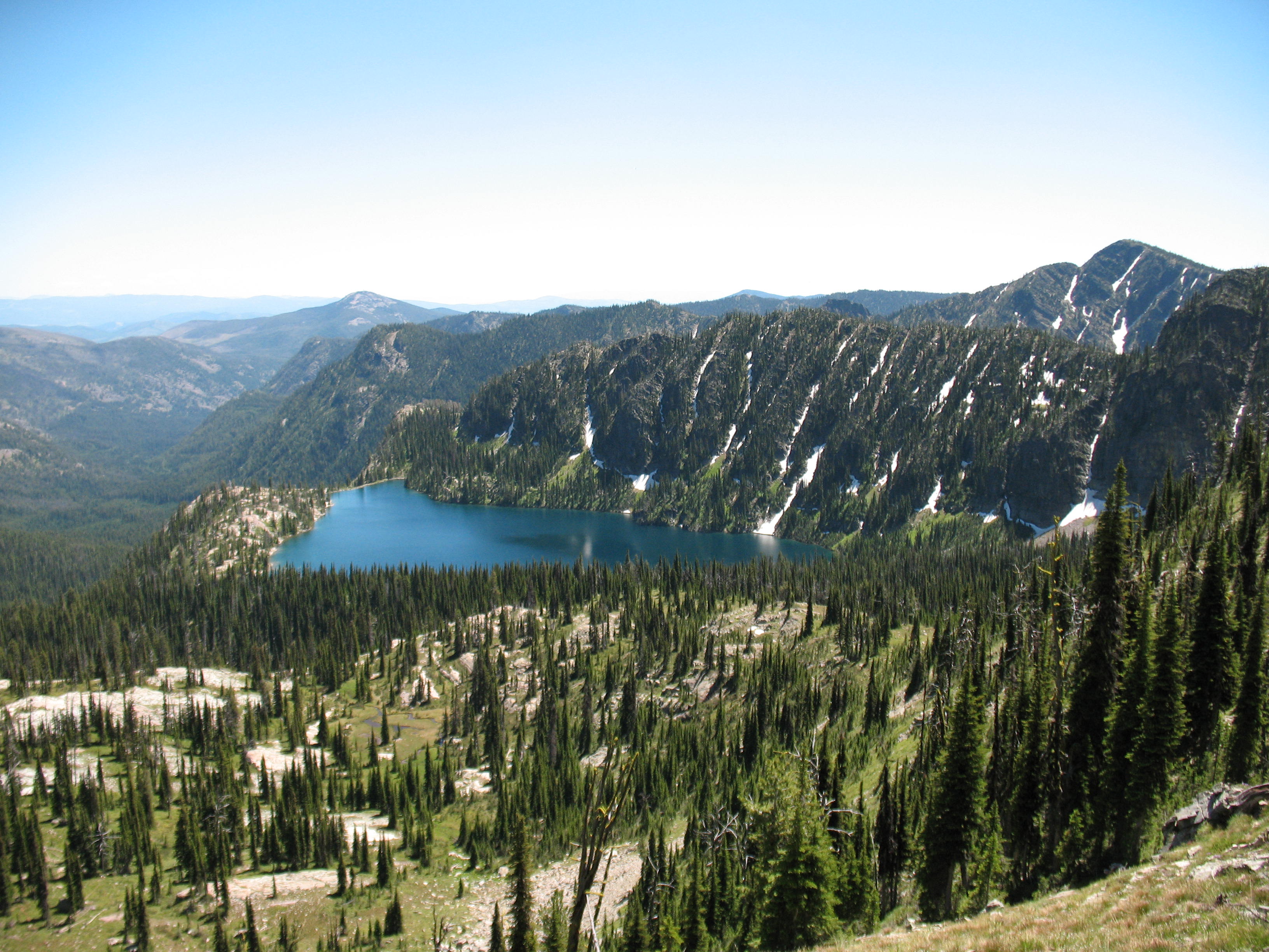 Alpine lake reflects blue sky, surrounded by mountain peaks with dusting of snow.