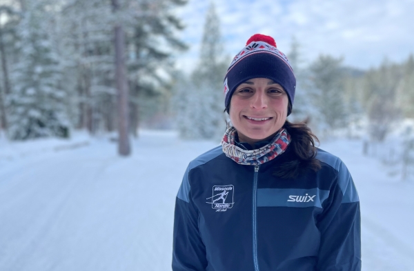 Young woman wearing a ski hat smiles at the camera, against a snowy, wooded background.