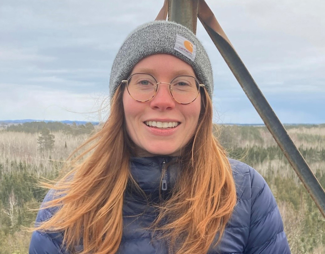 Woman in a stocking cap and glasses smiles at the camera while standing in an expansive prairie environment.