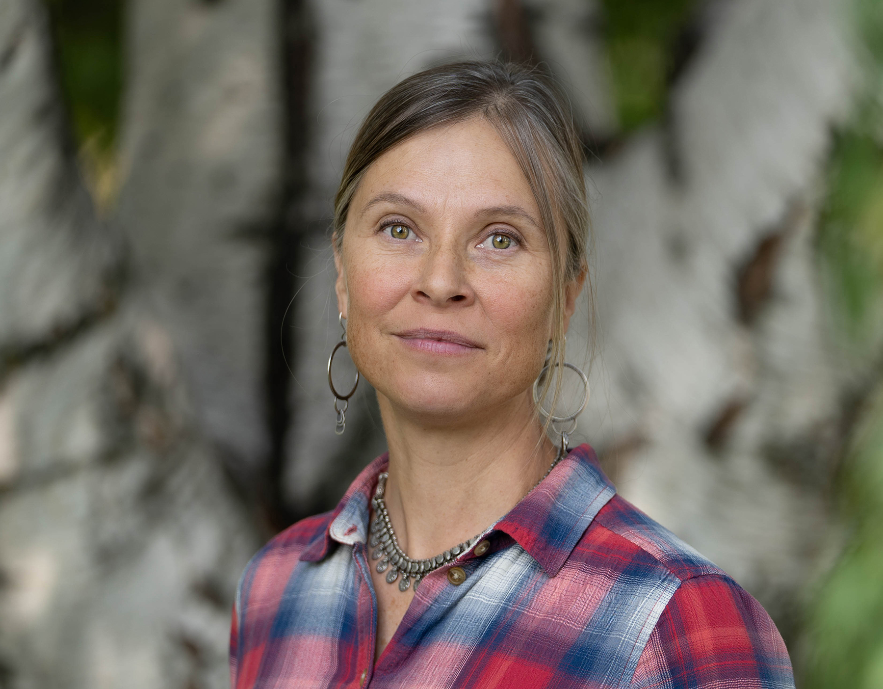 Woman with brown hair and green eyes, wearing a plaid shirt, smiles faintly at camera.